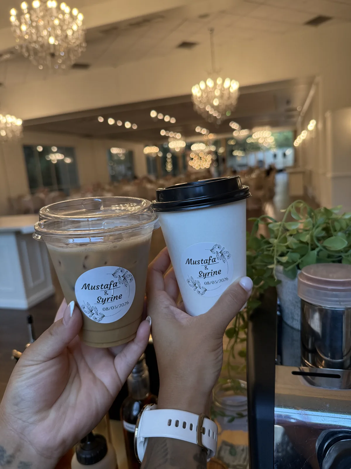 Iced latte and hot coffee in cups with a custom couple's-name wedding label, held up in a chandelier-lit DFW reception hall
